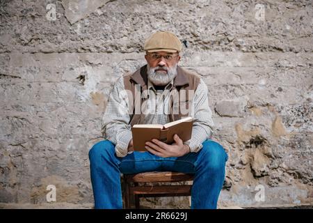 Un homme avec une barbe grise lit un livre contre un mur de béton Banque D'Images