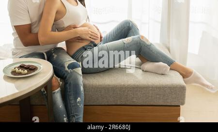 Un couple attendu s'assoit côte à côte sur le lit, le mari reposant ses mains avec amour sur le ventre de la femme enceinte Banque D'Images