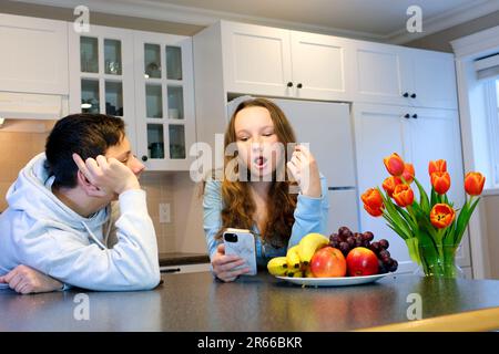 les adolescents dans la cuisine le garçon regarde une fille avec des yeux aimant une jeune femme mange des fruits de raisin un homme s'appuie sur la table Banque D'Images