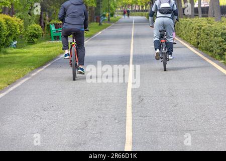 Deux jeunes hommes pédalent sur un chemin asphalté au milieu des arbres verts d'un parc de la ville par une journée ensoleillée. Copier l'espace. Banque D'Images