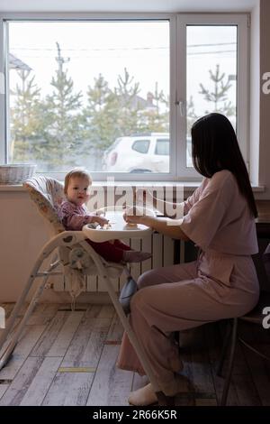 La jeune mère nourrit son bébé. Enfant assis à la table des enfants Banque D'Images