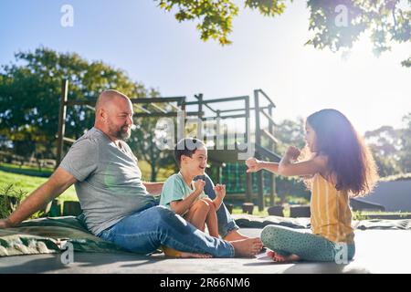 Père et enfants assis sur un trampoline dans une cour ensoleillée Banque D'Images