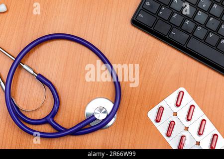 Vue à grand angle d'une table en bois où sont un stéthoscope, des pilules rouges sur une plaquette et un clavier d'ordinateur noir. Banque D'Images