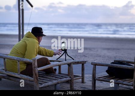 Un homme dans une veste jaune dans un café de plage s'assoit sur une chaise en bois et prend un coucher de soleil en mer sur un smartphone sur un trépied Banque D'Images