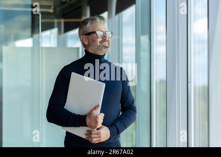 Portrait d'un vieil homme beau gris-cheveux designer, architecte, ingénieur debout dans le bureau, regardant par la fenêtre. Il tient un ordinateur portable entre ses mains, attendant une réunion d'affaires. Banque D'Images