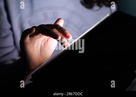 Gros plan d'une main d'une jeune fille méconnue touchant un écran de tablette numérique dans une pièce sombre. Banque D'Images