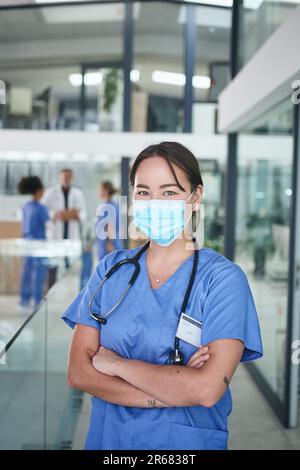 Je suis au plus heureux dans mes exfoliations. Portrait court d'une jeune infirmière attirante portant un masque facial et debout dans la clinique avec ses bras pliés. Banque D'Images