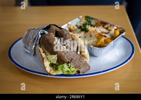 Un gyro méditerranéen grec enveloppé dans du papier d'aluminium sur une assiette avec un côté de frites grecques enrobées de mozzarella fondue et de fromage feta, d'herbes et d'épices Banque D'Images