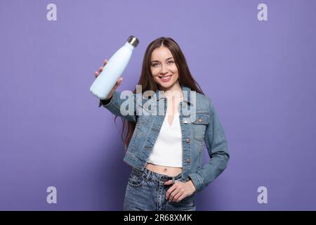 Belle jeune femme avec bouteille thermos sur fond violet Banque D'Images