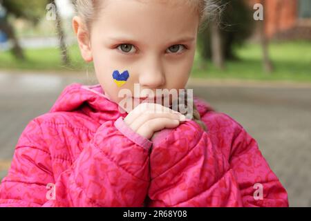 Petite fille avec dessin de drapeau ukrainien sur le visage en forme de coeur à l'extérieur, gros plan Banque D'Images