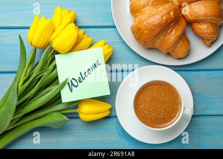 Carte de bienvenue, superbes tulipes jaunes, croissants et tasse de café aromatique sur table en bois bleu clair, plat Banque D'Images