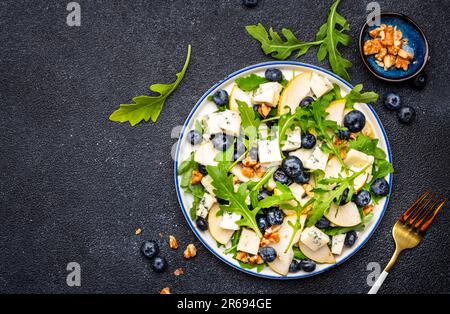 Salade gastronomique aux poires sucrées, aux bleuets, au fromage bleu, à l'arugula et aux noix. Arrière-plan de table noir, vue du dessus Banque D'Images