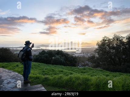 Tourisme prenant des photos avec un smartphone au sommet du Mont Eden au lever du soleil. Rangitoto Island au loin. Auckland. Banque D'Images