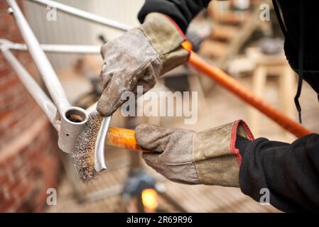 Travailleur méconnaissable avec des vêtements de travail, y compris des gants de protection à l'aide d'une brosse métallique au-dessus d'un cadre de vélo après avoir passé un chalumeau dessus pour le retirer Banque D'Images