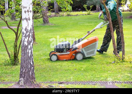 Le jardinier tond la pelouse et s'en occupe avec une tondeuse à essence dans le jardin près des arbres. Copier l'espace. Banque D'Images