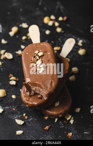 Pop-sutes au chocolat au lait et aux noisettes. Gros plan. Crèmes glacées, crèmes glacées, papillotes recouvertes de chocolat, bâtonnets, fond de pierre noire. Mise au point sélective. Banque D'Images