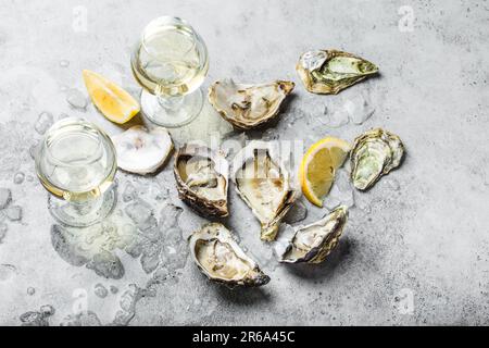 Gros plan d'une demi-douzaine d'huîtres fraîches ouvertes et de coquillages avec des quartiers de citron, deux verres de vin blanc ou de champagne, vue de dessus, béton rustique gris Banque D'Images