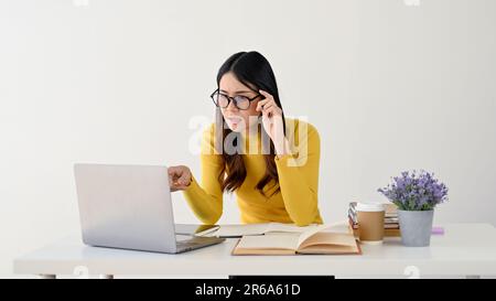 Une jeune étudiante asiatique perplexe et confuse touche ses lunettes, regarde son écran d'ordinateur portable avec hésitation et s'assoit à son de Banque D'Images