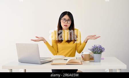 Une jeune asiatique curieuse et douée étudiante à l'université ou employée de bureau en lunettes se trouve à son bureau, haussant les épaules avec hésitation. ne le faites pas Banque D'Images