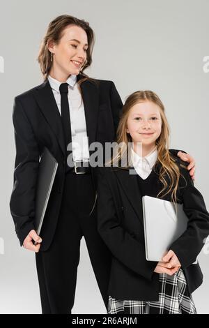 mère et fille, nomadisme numérique, travail à distance, e-learning, femme d'affaires en costume et fille debout avec des ordinateurs portables sur fond gris en st Banque D'Images