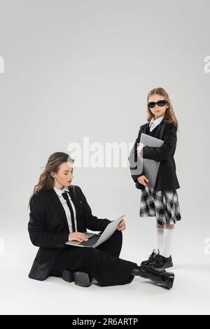 nomadisme numérique, femme tendance en costume utilisant un ordinateur portable près de fille dans l'uniforme d'école et des lunettes de soleil sur fond gris, travail à distance, mothe de travail Banque D'Images