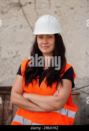 Jeune femme en casque blanc et gilet orange haute visibilité, cheveux longs foncés, mains croisées, en confiance. Rétrogr. Mur site de construction flou Banque D'Images
