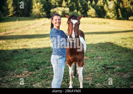 Femme en Jean et chemise debout à côté de brun et blanc pinto cheval foal, vert prairie derrière elle, photo de gros plan Banque D'Images