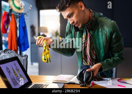 Créateur de mode transgenre biracial utilisant un ordinateur portable, parlant sur un smartphone et tenant le tissu Banque D'Images