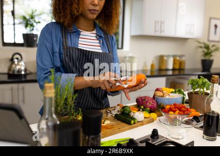 Femme biraciale ciblée préparant des légumes dans la cuisine épluchant la carotte Banque D'Images