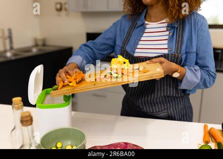 Section médiane de la femme biraciale en tablier compostage de déchets végétaux dans la cuisine Banque D'Images