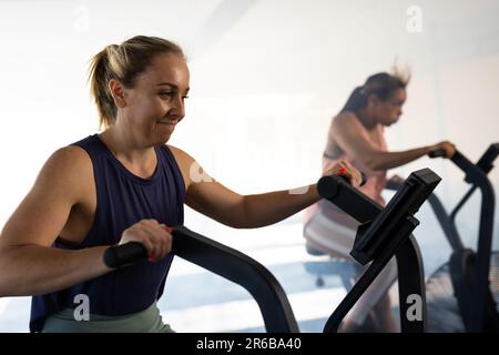 Souriante jeune femme caucasienne s'exerçant avec confiance sur machine elliptique d'entraînement dans la salle de gym Banque D'Images