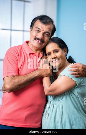 Prise de vue verticale d'un heureux couple indien senior debout en s'embrassant tout en regardant la caméra à la maison - concept de la relation familiale Banque D'Images