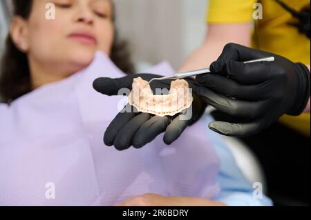 Gros plan les mains gantées d'orthodontiste tenant le moule en plâtre dentaire sur un arrière-plan flou d'un patient dans la chaise du dentiste Banque D'Images