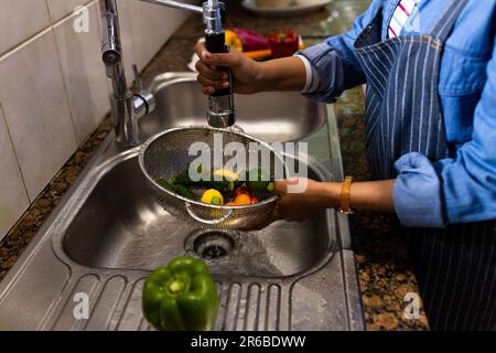 Section médiane de la femme biraciale en tablier lavant la passoire de légumes dans l'évier de cuisine Banque D'Images