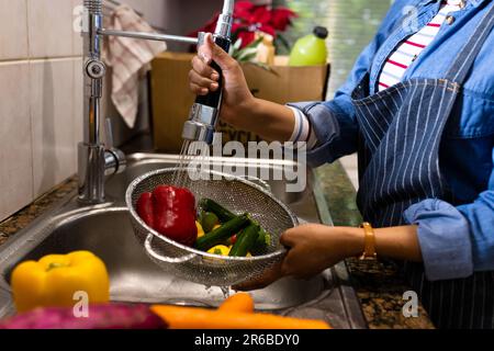 Section médiane de la femme biraciale en tablier lavant la passoire de légumes dans l'évier de cuisine Banque D'Images