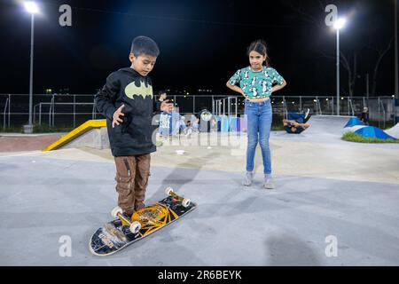 San Salvador, San Salvador, El Salvador - 11 novembre 2022 : deux jeunes enfants bruns jouent avec joie avec un skateboard dans un parc de skate la nuit contre le brig Banque D'Images