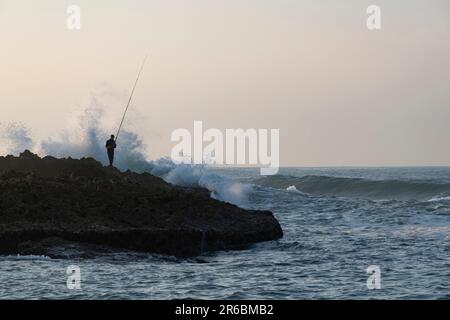 Une silhouette de pêcheur debout sur un affleurement rocheux de l'océan Atlantique avec sa canne à pêche pointait vers les vagues qui se brisent à Oualidia, au Maroc Banque D'Images