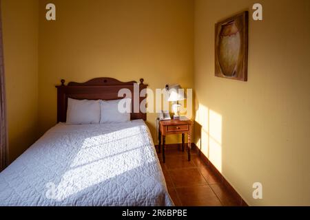 San Salvador, San Salvador, El Salvador - petite chambre avec un lit avec draps et oreillers blancs, téléphone, lampe et une peinture sur les murs jaunes dedans Banque D'Images