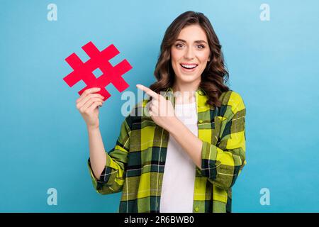 Photo de la jolie dame positive indiquer le doigt main tenir le symbole de hashtag de papier isolé sur fond bleu Banque D'Images