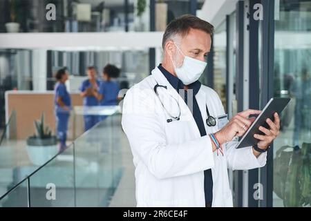 Nous avons passé une journée bien remplie. un beau médecin mature debout et portant un masque facial tout en utilisant une tablette numérique. Banque D'Images