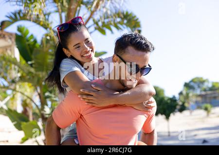 Souriant jeune ami caucasien coggydorant petite amie tout en se tenant à la plage contre le ciel clair. Copier l'espace, non modifié, amour, ensemble, vacances, Banque D'Images
