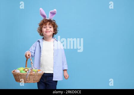 Joyeux garçon dans un joli bandeau d'oreilles de lapin tenant un panier en osier avec des œufs de Pâques sur fond bleu clair. Espace pour le texte Banque D'Images