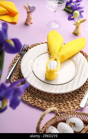 Table festive avec œufs peints, assiettes et fleurs d'iris sur fond lilas. Fête de Pâques Banque D'Images