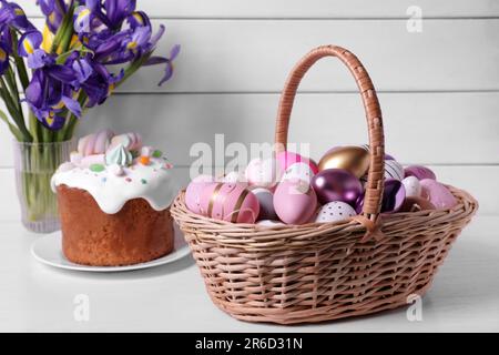 Panier en osier avec œufs décorés avec soin, gâteau de Pâques et fleurs d'iris sur table blanche Banque D'Images