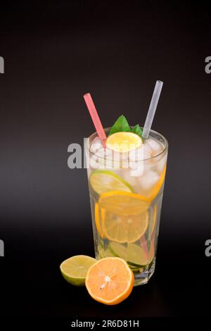 Un grand verre d'un mélange de jus d'agrumes avec de la glace, des pailles et des feuilles sur fond noir, à côté de tranches de citron et de lime. Gros plan. Banque D'Images