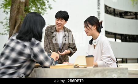 Un jeune étudiant asiatique élégant et gai discute de travail avec ses amis tout en travaillant sur leur projet de groupe au parc de campus toge Banque D'Images