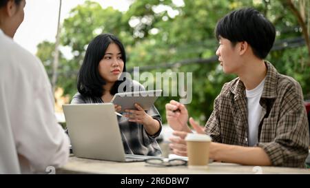 Une jeune étudiante asiatique sérieuse discutant du travail et partageant ses idées avec ses amis tout en travaillant sur leur projet de groupe à une table dans Banque D'Images