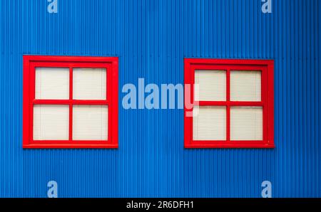 Deux fenêtres avec cadres en bois rouge dans un mur bleu. Extérieur d'une maison avec fenêtres et stores sont fermés. Belle façade de maison colorée. Personne Banque D'Images