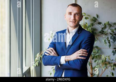Un homme d'affaires en costume bleu marine et cravate assortie se tient en toute confiance devant une grande fenêtre, en regardant au loin Banque D'Images