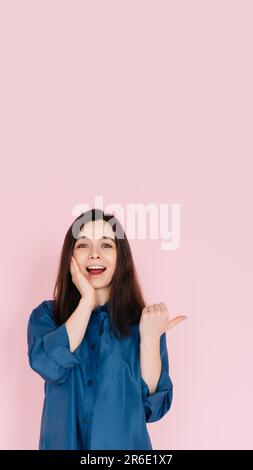 Jeune femme exprimant étonnement et joie, touchant la joue et pointant le doigt sur l'espace vide, avec un grand sourire crasseux sur fond rose isolé Banque D'Images